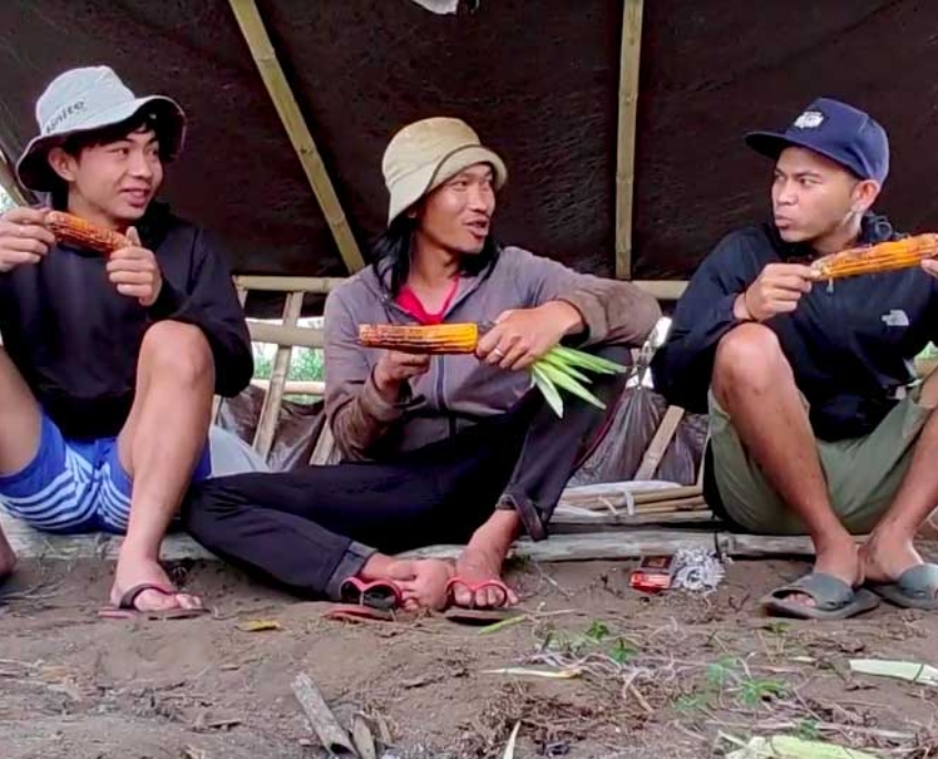 tres jóvenes indígenas sentados juntos bajo un refugio, compartiendo maíz asado mientras sonríen y charlan