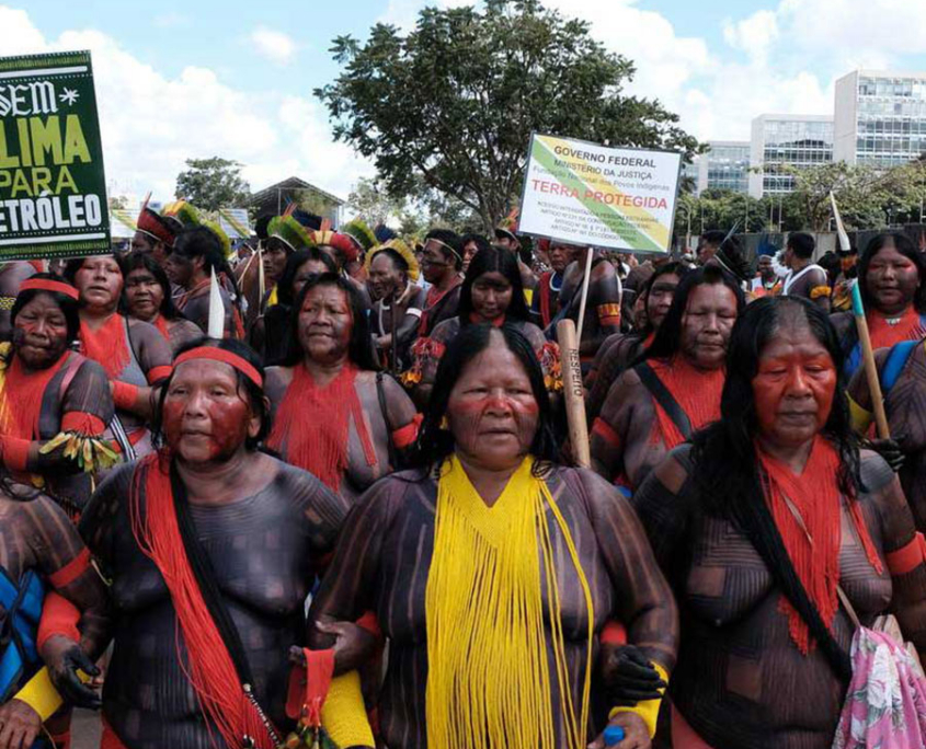 large group of Indigenous women from Brazil in traditional attire protest with signs, advocating for climate action and land protection