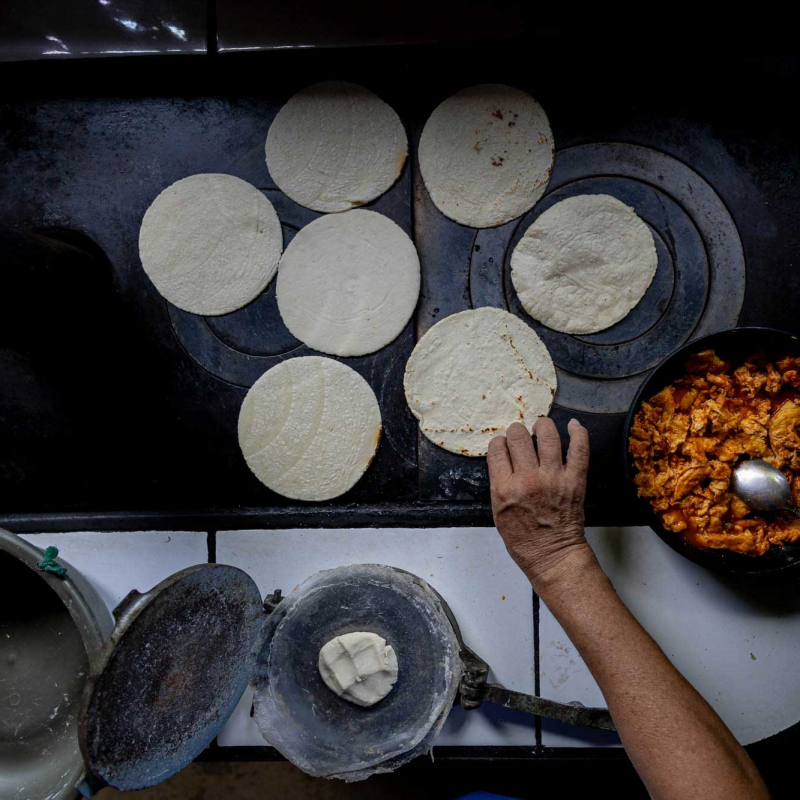 una mano voltea tortillas en un hornillo, con una sartén de relleno cocido y una prensa de masa cerca, en una cocina tradicional
